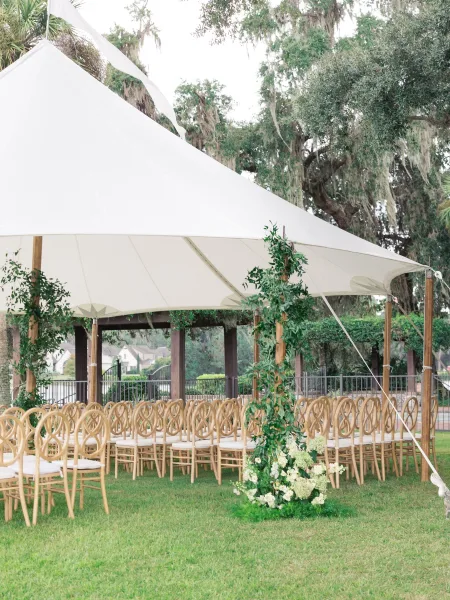 Outdoor ceremony setup with sailcloth tent wedding seating, wooden chairs lining an aisle with greenery garland and floral ground arrangement on a lawn