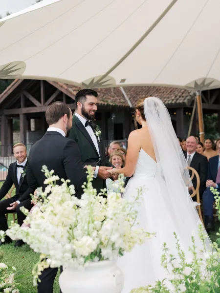 Wedding vows as bride and groom hold hands, her bridal veil flowing, at an outdoor lawn ceremony with guests under a canopy tent