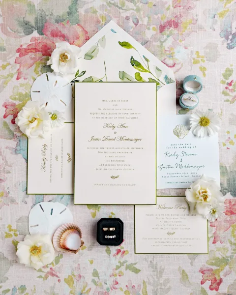 Invitation suite wedding invitation flat lay with gold foil calligraphy, shell illustration, rings in boxes, and seashells on floral linen backdrop