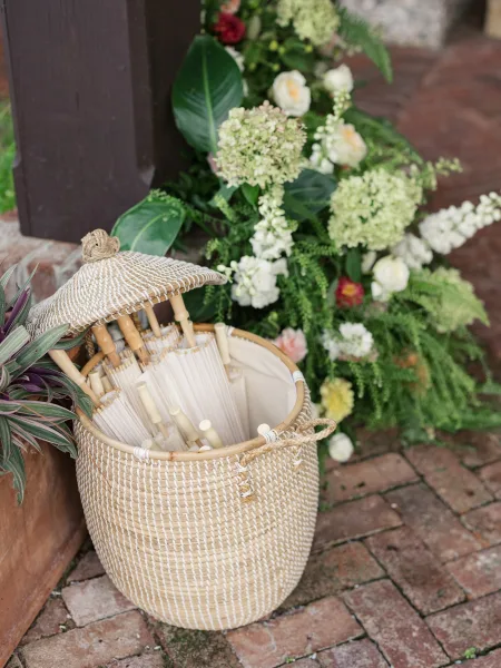 Wedding parasol basket filled with white paper parasols and tropical greenery, set beside florals on a brick patio near a porch column