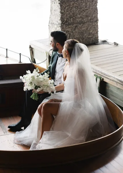 Couple portrait of bride and groom sitting on a wooden boat, her long veil draping as they hold a white bouquet by the dock and water