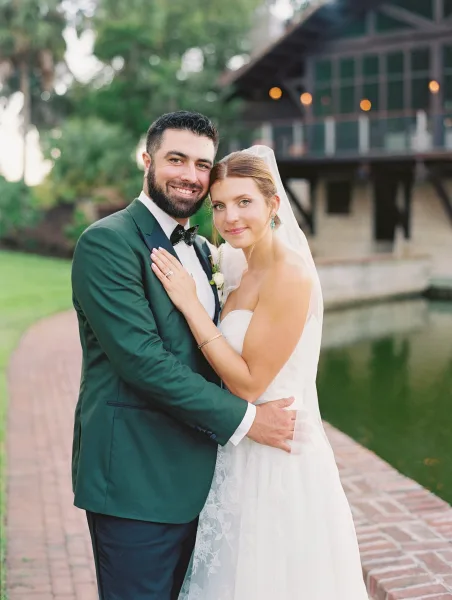 Couple portrait of bride and groom hugging, her hand on his chest, veil and green tux by a brick path with pond and string lights