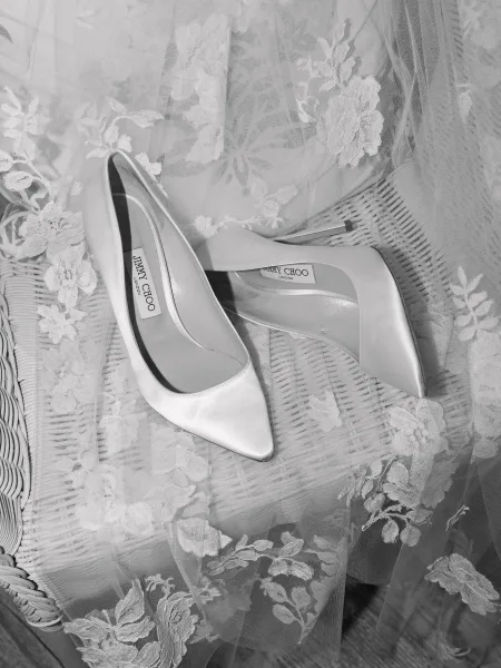 Bridal shoes with white pointed toe heels resting on a lace veil accent, styled on a wicker chair over a warm wood floor