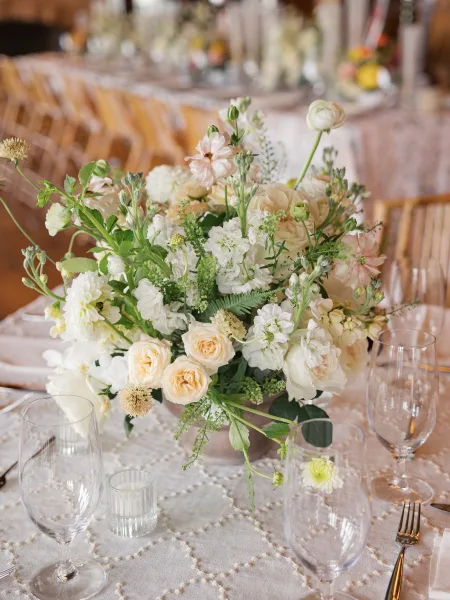 Wedding centerpiece with white flowers and blush blooms in a ceramic vase, styled with greenery, gold flatware, glasses, and candlelight on patterned linen tables at a reception