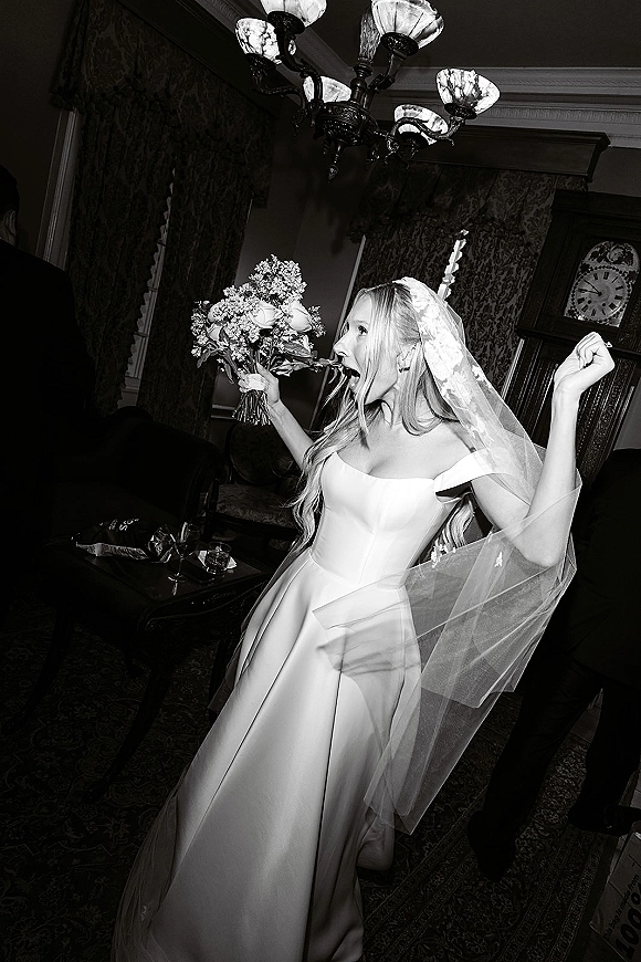 Bridal portrait in a black and white wedding photo of a bride holding bouquet, laughing in a vintage room with chandelier and curtains