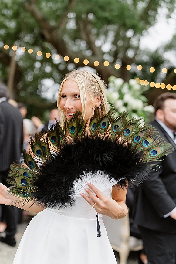 Bride portrait holding a peacock feather fan in a strapless wedding dress, smiling at an outdoor reception under bistro lights