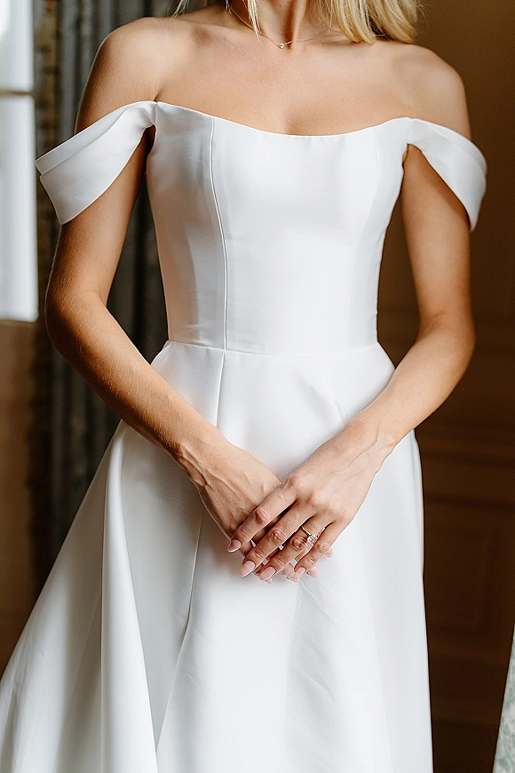 Wedding dress with off the shoulder sleeves in smooth satin, showing seam detailing, necklace and engagement ring by window light