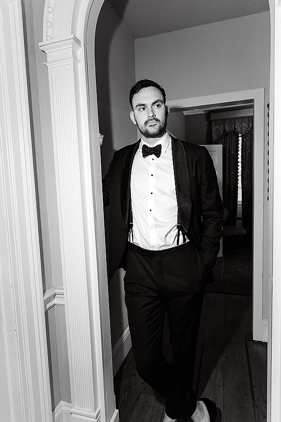Groom portrait in a black tie tuxedo with bow tie and suspenders, leaning in an interior doorway with hallway curtains and wood floor