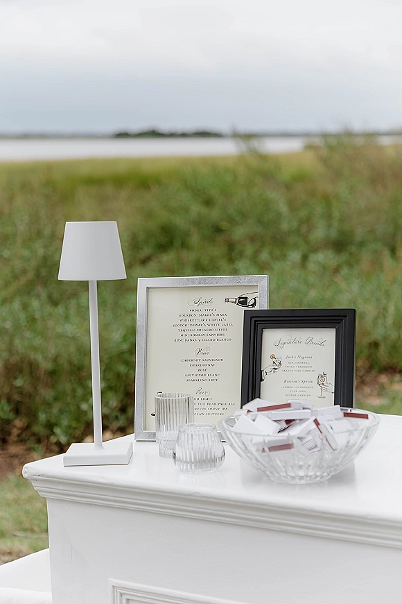 Wedding bar signage with framed drink menus, a white table lamp, and escort cards in a glass bowl set by a waterfront field under open sky