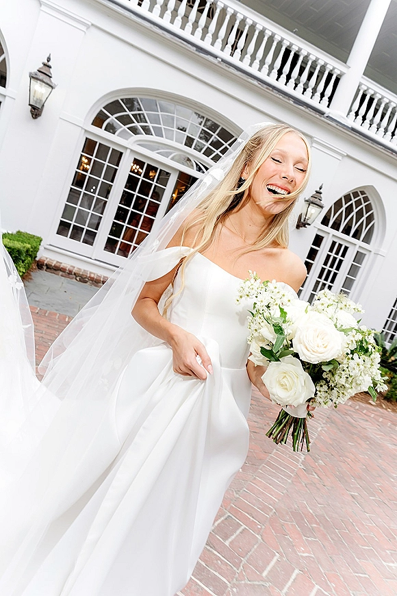 Bridal portrait of a laughing bride holding bouquet with a long veil, strapless dress, and white rose blooms outside a white facade