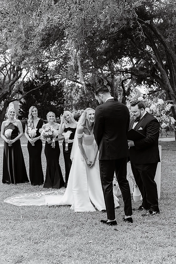 Wedding ceremony moment with bride and groom at altar, bride in veil smiling as officiant reads vows under trees with moss on lawn