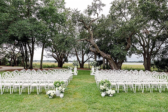 Outdoor ceremony setup with wedding aisle decor, white chairs in rows lining a grass aisle with pedestal florals under oak trees by water