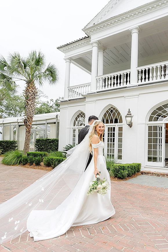 Bride and groom portrait with the bride looking back, long veil trailing and bouquet in hand on a brick courtyard by white columns