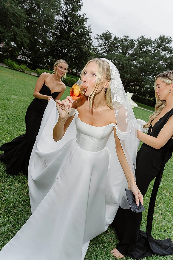 Bride portrait of her drinking champagne from a flute while bridesmaids adjust her veil on a grassy lawn under an overcast sky