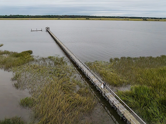 Couple portrait from a drone wedding photo of bride in white gown and groom in dark suit on a wooden dock amid marsh grass and water