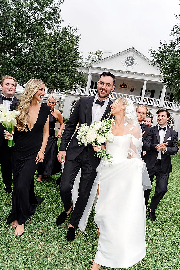 Wedding party portrait with bride holding a white bouquet and long veil, bridesmaids in black dresses and tuxedos before a white mansion facade