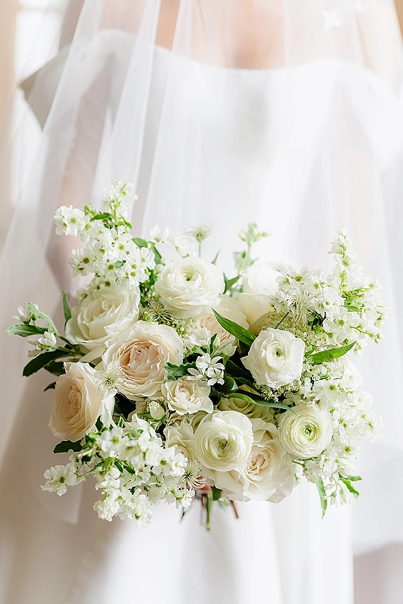 Bridal bouquet of white roses and ranunculus with baby’s breath and greenery, held against a bridal gown and veil in soft indoor light