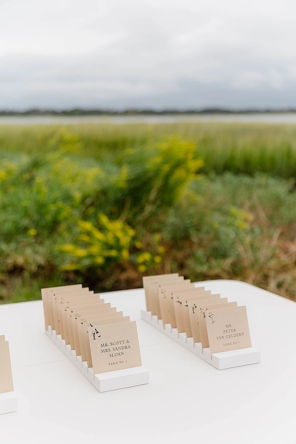 Wedding place cards arranged in white holders on a white table, escort card display set outdoors by wildflowers and water under cloudy sky