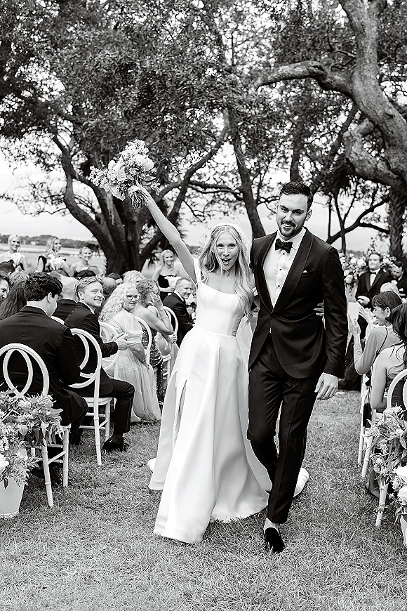 Wedding recessional as bride and groom walking aisle hand in hand, bride raising bouquet past guests on a waterfront lawn beneath trees