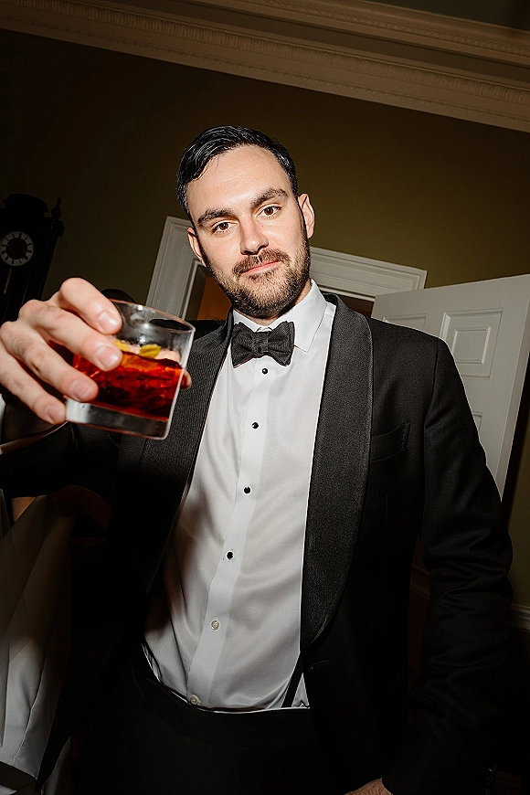 Groom portrait in a black tuxedo holding a cocktail glass, bow tie and suspenders visible, standing by a doorway in an indoor room