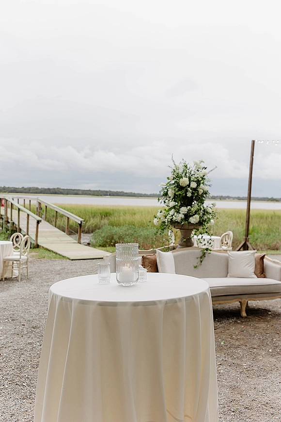 Wedding lounge area with outdoor wedding lounge sofa, round cocktail table and candles under string lights by a waterfront dock with marsh grass