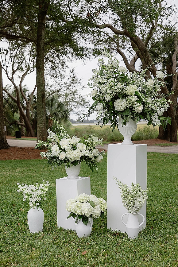Ceremony floral arrangements with wedding aisle floral urns on white pedestals, featuring white roses, hydrangeas, and greenery by a marsh lawn path