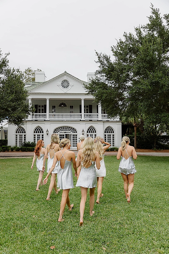 Bridesmaids getting ready in matching satin slip dresses walk away barefoot across a lawn toward a white mansion under overcast skies