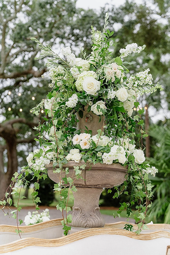 Wedding floral arrangement in a stone urn with white roses, greenery, and trailing ivy, set outdoors beneath trees and string lights