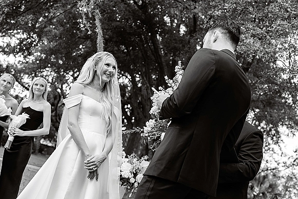 Wedding vows as groom reads to smiling bride holding hands, her bridal veil flowing amid bridesmaids and floral altar under trees