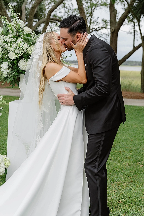Wedding kiss portrait of bride and groom kissing under a white floral arch by a lakeside lawn, lace veil draped over his black tuxedo