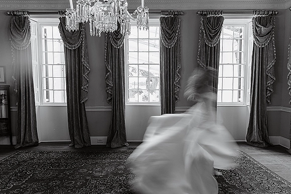 Bride portrait in black and white, twirling in a flowing wedding dress with soft motion blur beneath a chandelier by tall windows