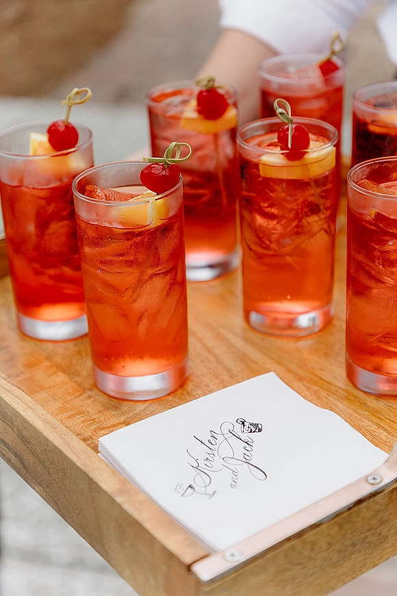 Wedding signature cocktail in highball glasses, pink and iced with citrus slices and cherries, on a wooden tray over neutral fabric tabletop