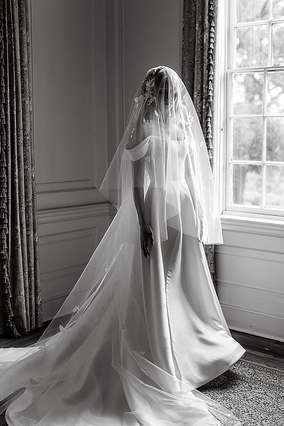 Bridal portrait in black and white of a bride in an off-the-shoulder gown with cathedral veil over her face by a tall window