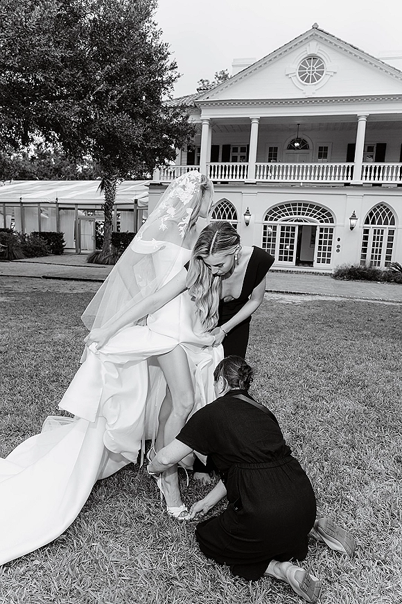 Bridal getting ready as a bridesmaid helps the bride putting on shoes, adjusting her dress and veil on a lawn by a white mansion porch