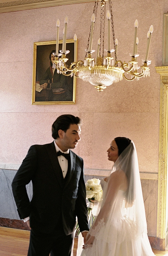Couple portrait of bride and groom holding hands, gazing at each other beneath a chandelier in a pink, ornate hallway with bouquet and veil
