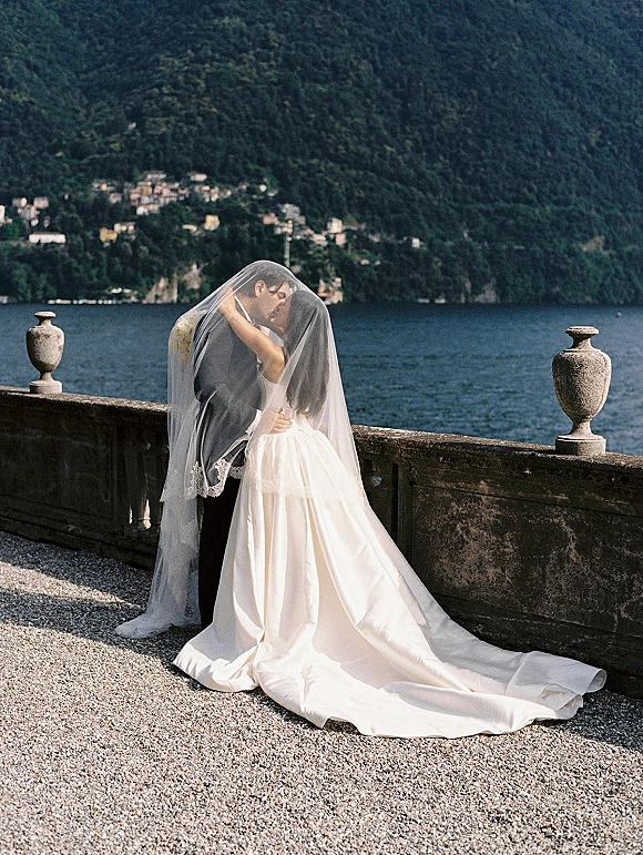 Wedding kiss as bride and groom embrace under a lace-trim veil on a stone terrace with balustrade, lake and mountains behind