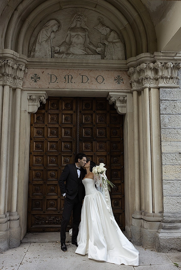Wedding kiss portrait of bride and groom kissing in a church doorway, bride in strapless gown and veil holding a white rose bouquet