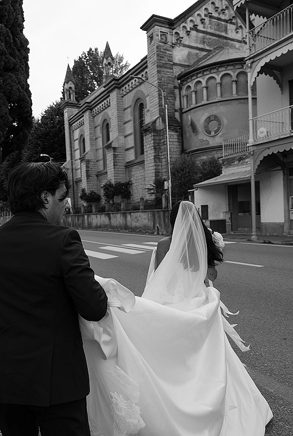 Wedding couple walking away across a city crosswalk, groom holding dress train as bride’s long veil flows behind her in a strapless gown