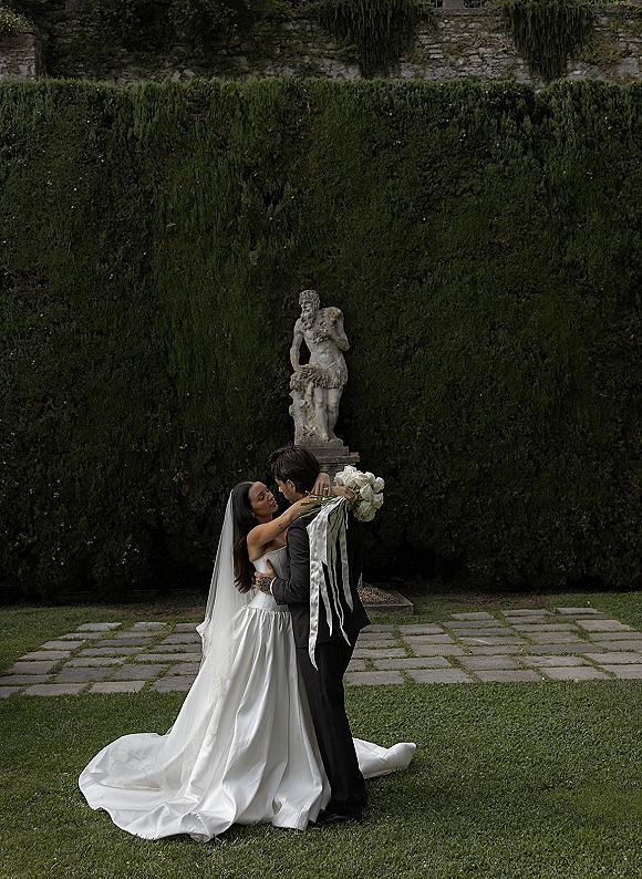 Couple portrait of bride and groom embrace, bride holding a white rose bouquet with veil in a formal garden by a stone statue