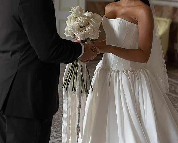 Wedding vows as bride and groom hold hands during ring exchange, her strapless dress and veil beside a white rose bouquet on patterned carpet