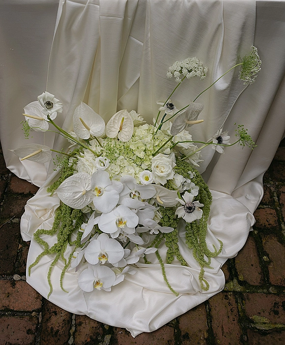 Bridal bouquet with white orchid blooms cascading over roses and anemones, styled on draped satin fabric atop a brick floor