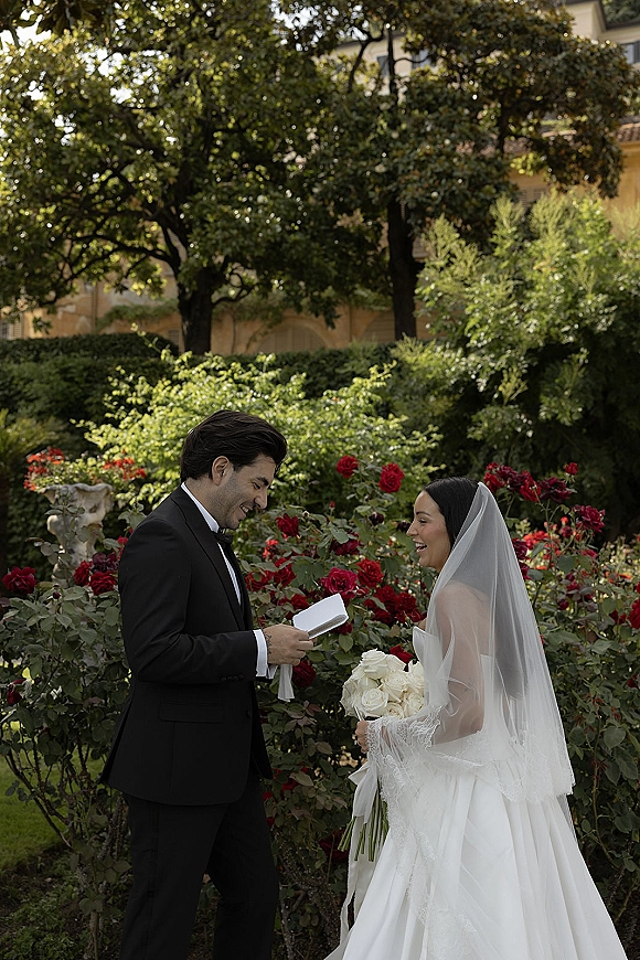 Wedding vows as groom reads from a vow booklet to his bride in a rose garden, her long veil and white rose bouquet in hand