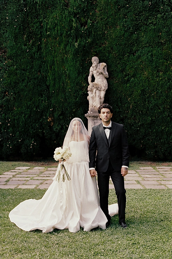 Couple portrait of bride and groom holding hands, bride in strapless dress with veil and ribbon bouquet beside a garden statue