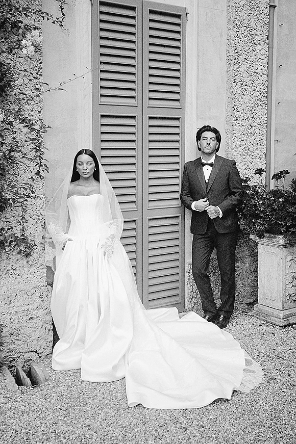 Couple portrait in black and white, bride in strapless gown with long train and veil beside groom in tuxedo by ivy stucco wall