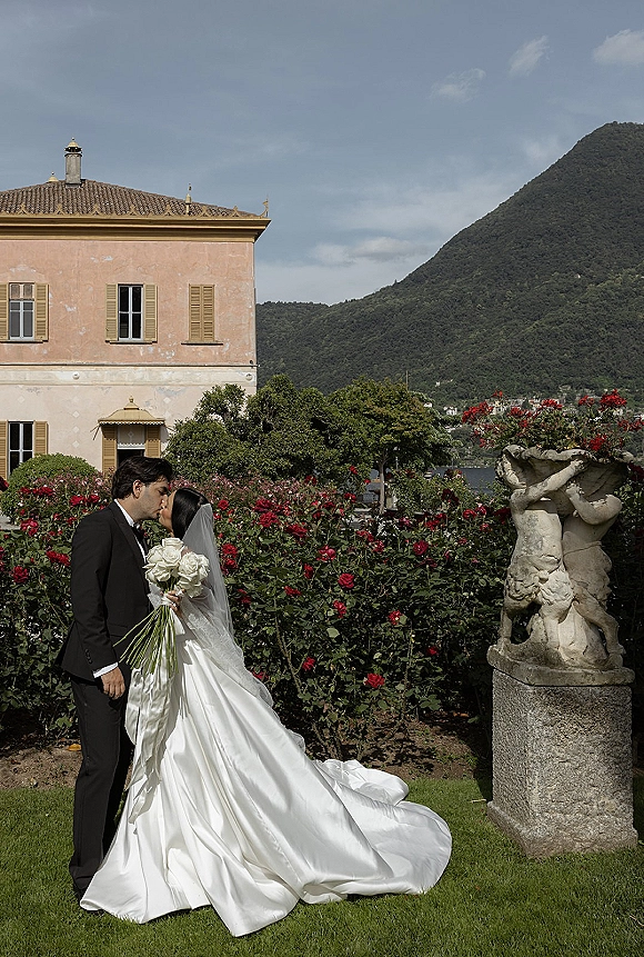 Wedding kiss portrait of bride and groom kiss, her long veil and white rose bouquet beside a villa garden with lake and mountains