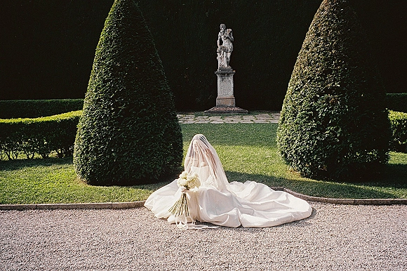 Bridal portrait of a bride sitting with cathedral veil and dress train spread, holding a white rose bouquet with ribbon in a formal topiary garden