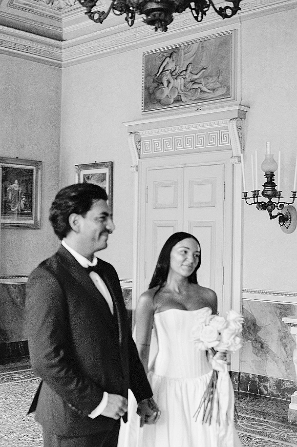 Couple portrait in a black and white wedding portrait style, bride with bouquet and groom in tuxedo holding hands in ornate interior under chandelier