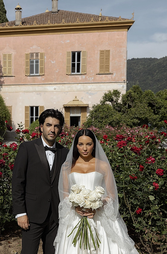 Couple portrait of bride in strapless dress with veil holding a white rose bouquet beside groom in black tux, in a rose garden by a villa facade