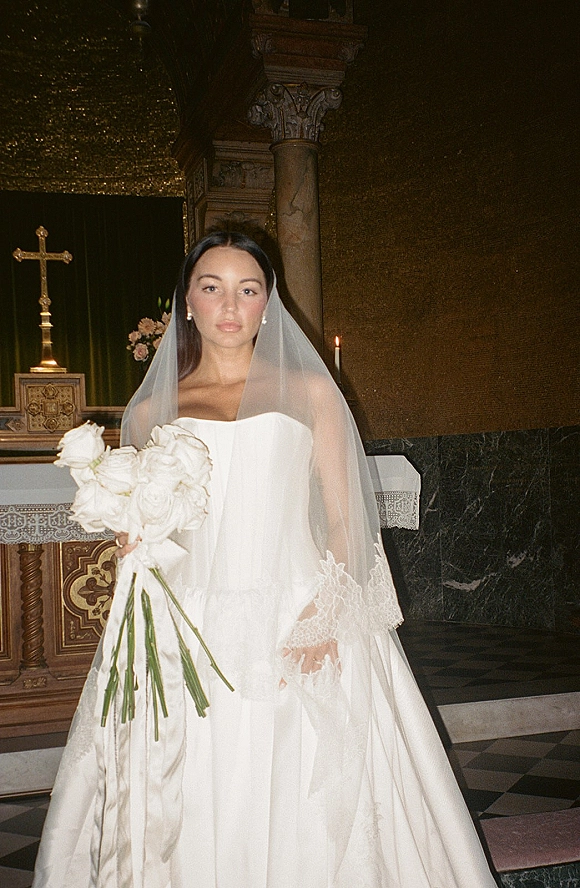 Bridal portrait of a bride in a strapless wedding dress with cathedral veil, holding a white rose bouquet at a church altar with cross