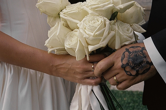 Wedding bouquet of white roses with long ribbon held by bride and groom hands, wedding rings visible against blurred garden greenery